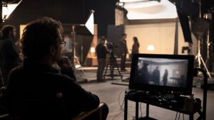 Director on a soundstage watching actors rehearse near a cinema camera and blurred monitor references